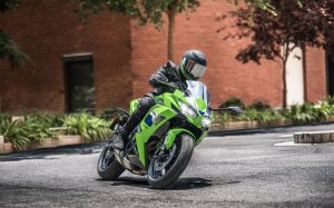 Kawasaki Ninja 650 2027 in Lime Green in Schräglage in einer Kurve vor einer roten Backsteinwand, Fahrer mit grün-schwarzem Helm