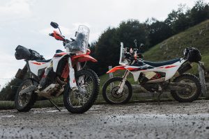 KTM 690 Rally 2027 – zwei verschmutzte Maschinen mit Gepäckrollen auf nassem Schotterplatz vor bewaldetem Hang
