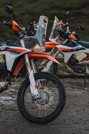 KTM 690 Rally 2027 – Nahaufnahme der Front zweier nasser Maschinen mit Windschild, WP-Gabel und Speichenvorderrad auf Schotterplatz