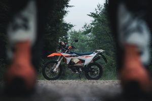 KTM 690 Rally 2027 – Seitenansicht von rechts auf Waldweg, gerahmt von zwei unscharfen Motorrädern im Vordergrund