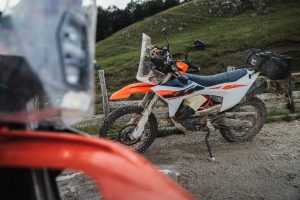 KTM 690 Rally 2027 – verschmutzte Maschine mit Gepäckrolle auf dem Heckträger an einem Schotterparkplatz in den Bergen