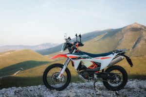KTM 690 Rally 2027 – Seitenansicht von rechts auf Steinuntergrund mit Blick über grüne Bergkuppen und Schotterpiste