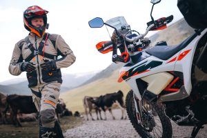 KTM 690 Rally 2027 – Fahrer in Enduroausrüstung neben der Maschine auf Schotterpiste, Kuhherde und Berghänge im Hintergrund