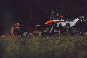 KTM 690 Rally 2027 – Seitenansicht auf einer Waldlichtung, zwei Männer entfachen daneben ein Lagerfeuer