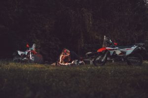 KTM 690 Rally 2027 – zwei Maschinen links und rechts eines Lagerfeuers am Waldrand in der Abenddämmerung