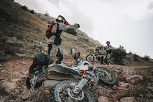 KTM 690 Rally 2027 – abgelegte Maschine mit Gepäckrolle im Geröll eines steilen Hangs, zwei Fahrer daneben