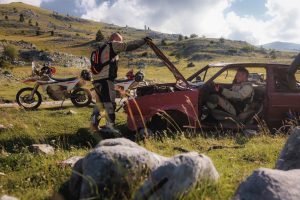 KTM 690 Rally 2027 – zwei Maschinen auf einer Bergwiese abgestellt, Fahrer an einem ausgeschlachteten Autowrack