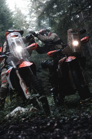 KTM 690 Rally 2027 – zwei schlammverkrustete Maschinen frontal im Wald, Scheinwerfer an, Fahrer dahinter