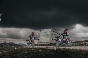 KTM 690 Rally 2027 – zwei Fahrer mit Gepäck auf einer Hochebene vor einer Ruine unter dunkler Gewitterwolke