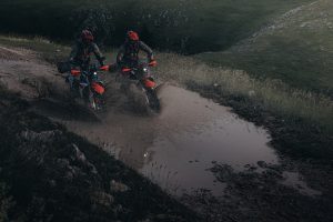 KTM 690 Rally 2027 – zwei Fahrer durchqueren nebeneinander eine schlammige Wasserpassage in grüner Berglandschaft