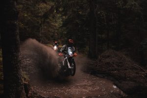 KTM 690 Rally 2027 – Wasserdurchfahrt auf dunklem Waldweg mit hoher Fontäne, zweiter Fahrer mit Licht im Hintergrund