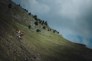 KTM 690 Rally 2027 – Wheelie im Stehen an einem steilen grünen Berghang, Totale vor bewölktem Himmel