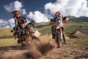 KTM 690 Rally 2027 – Heckansicht zweier Maschinen beim Anfahren auf Schotter, aufgewirbelte Steine vor Bergpanorama