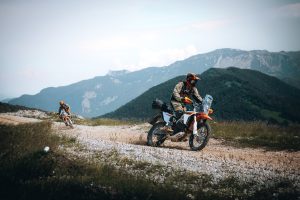KTM 690 Rally 2027 – Seitenansicht mit Fahrer und Gepäckrolle auf Schotterpiste, Bergkette im Dunst dahinter