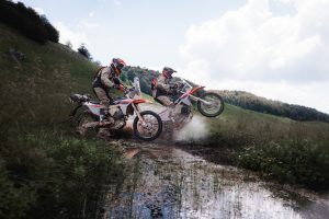 KTM 690 Rally 2027 – zwei Fahrer sprengen durch eine Wasserlache auf einer Bergwiese, eine Maschine mit angehobenem Vorderrad