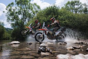 KTM 690 Rally 2027 – zwei Fahrer durchqueren mit den Rally-Enduros eine Flussfurt, das Wasser spritzt hoch auf