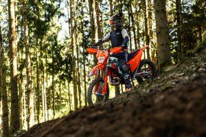 KTM 300 EXC 6DAYS 2027 – Fahrer mit angehobenem Vorderrad an einem Waldhang im Gegenlicht