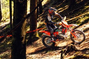 KTM 300 EXC 6DAYS 2027 – Fahrer in Schräglage auf einem staubigen Waldweg, Hinterrad wirft Erde auf