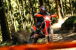 KTM 450 EXC-F 6DAYS 2027 – Fahrer in Schräglage mit eingeschaltetem Scheinwerfer und Staubfahne im Wald