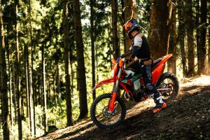 KTM 450 EXC-F 6DAYS 2027 – Fahrer fährt einen Anstieg durch dichten Nadelwald hinauf