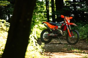 KTM 300 EXC 6DAYS 2027 – Zweitakt-Enduro auf einem Waldweg im gefleckten Sonnenlicht
