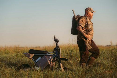 LiveWire S4 Honcho Trail 2026 – Jäger mit Gewehrtasche geht neben dem abgestellten Minibike im Marschgras bei Sonnenuntergang