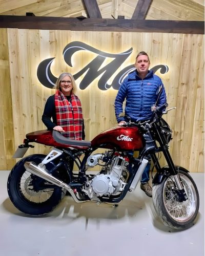 Mac Motorcycles Rex 2026 – Gründer Ian Oliver und Besucherin mit Mac Ruby in Flame Red im Mac Motorcycles HQ, beleuchtetes Mac-Logo an Holzwand