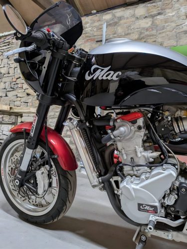 Mac Motorcycles Ruby 2026 – Frontansicht in Schwarz-Silber mit Café-Racer-Cockpitverkleidung in der Werkstatt-Scheune mit Natursteinwänden