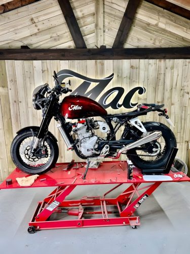 Mac Motorcycles Ruby 2026 – Linke Seitenansicht in Flame Red auf roter Werkstatthebebühne, Mac-Logo an Holzwand im Hintergrund