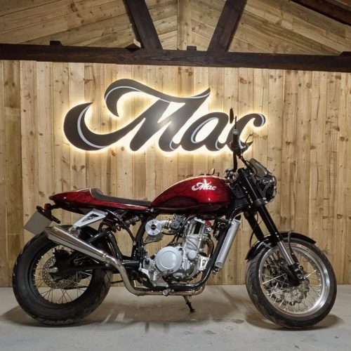 Mac Motorcycles Ruby 2026 – Komplette rechte Seitenansicht in Flame Red vor beleuchtetem Mac-Schriftzug, Showroom-Atmosphäre im HQ