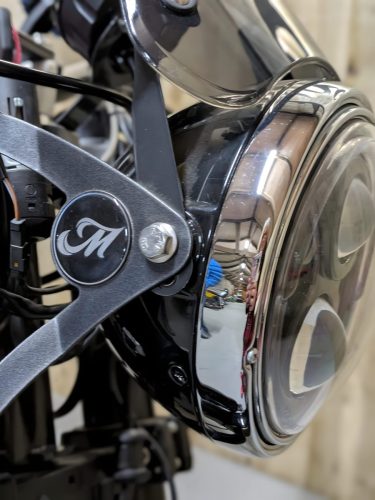Mac Motorcycles Rex Ruby 2026 – Detailaufnahme des runden Scheinwerfers mit Chromring und graviertem Mac-M-Badge an der Halterung