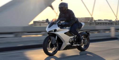 Norton Manx R Signature 2026 – Fahrer auf silberner Manx R in Schräglage auf Brücke bei Sonnenuntergang, Seitenansicht in Fahrt