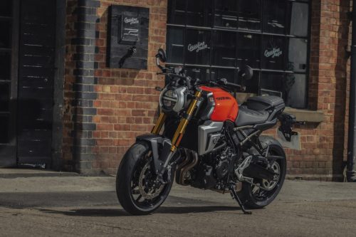 Suzuki GSX-8T 2027 in Glass Blaze Orange, Frontansicht mit Rundscheinwerfer vor Backsteinfassade mit Rundbogenfenstern