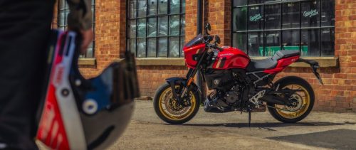 Suzuki GSX-8TT in Rot mit goldenen Felgen vor einer Backsteinhalle, im Vordergrund ein Helm