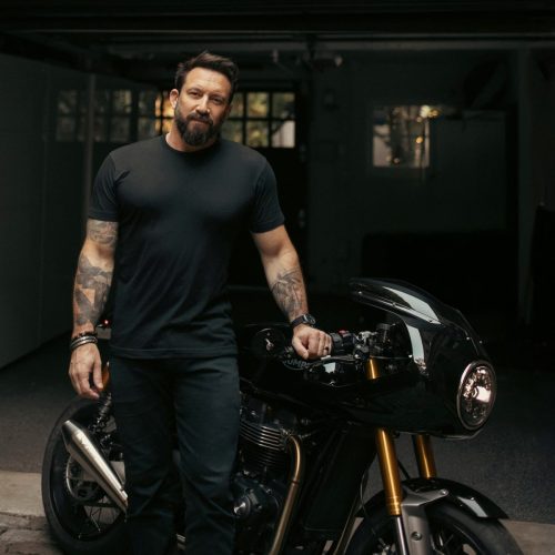 Eric Hendrikx in schwarzem Shirt neben einem schwarzen Triumph Café Racer mit Halbschale und goldener Gabel vor einer Garage