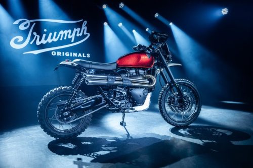 Triumph Scrambler 1200 Custom-Umbau aus Frankreich – Scrambler mit rotem Metallflake-Tank und Doppelauspuff im Scheinwerferlicht vor Triumph-Originals-Logo