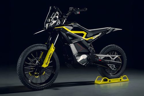 Ultraviolette Shockwave – Dreiviertelansicht von vorne links in Cosmic Black mit gelbem Gabelschutz, Rallye-Navigationsturm und Offroad-Bereifung auf gelbem Montageständer