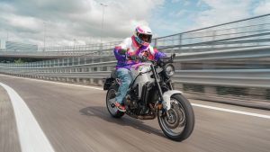 Yamaha XSR900 2027 – Fahraufnahme in Neon White auf einer Stadtautobahn mit Leitplanken, Fahrer in Retro-Jacke in Weiß, Pink und Blau