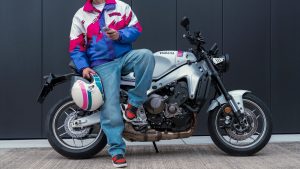 Yamaha XSR900 2027 – Fahrer in 90er-Jahre-Jacke lehnt mit Helm an der XSR900 in Neon White vor dunkler Wand