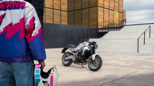 Yamaha XSR900 2027 – XSR900 in Neon White vor goldener Glasfassade und Treppe, im Vordergrund Fahrer mit Retro-Helm