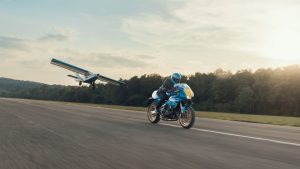 Yamaha XSR900 GP 2027 – Fahraufnahme in Legend Blue bei tiefstehender Sonne, im Hintergrund ein tief fliegendes Kleinflugzeug