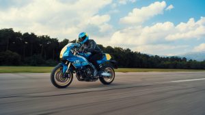 Yamaha XSR900 GP 2027 – Fahraufnahme in Legend Blue in der Seitenansicht vor Wald und Wolkenhimmel