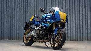 Yamaha XSR900 GP 2027 – schräge Frontansicht in Legend Blue mit goldenen Felgen vor einer Wellblechwand
