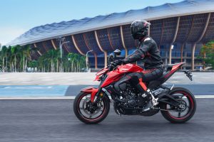 Zontes 703R 2027 - Lava Red, Fahraufnahme mit Fahrer in Schwarz vor einer modernen Stadionkulisse mit Palmen